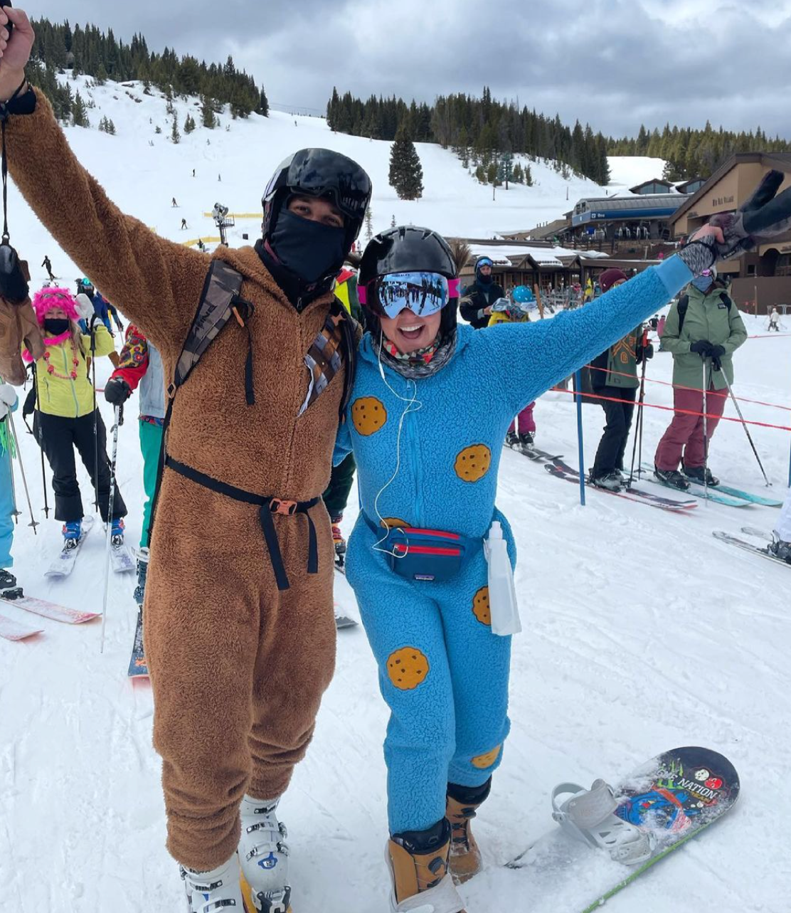 two people in costumes and helmets smiling with hands in air at a ski resort