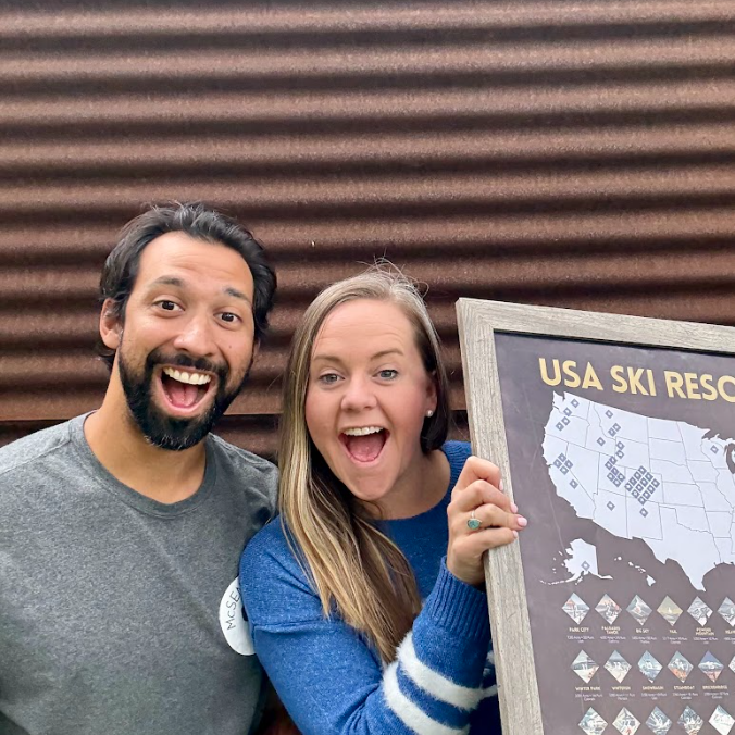 Two people posing with a framed map of USA ski resorts against a corrugated metal wall.