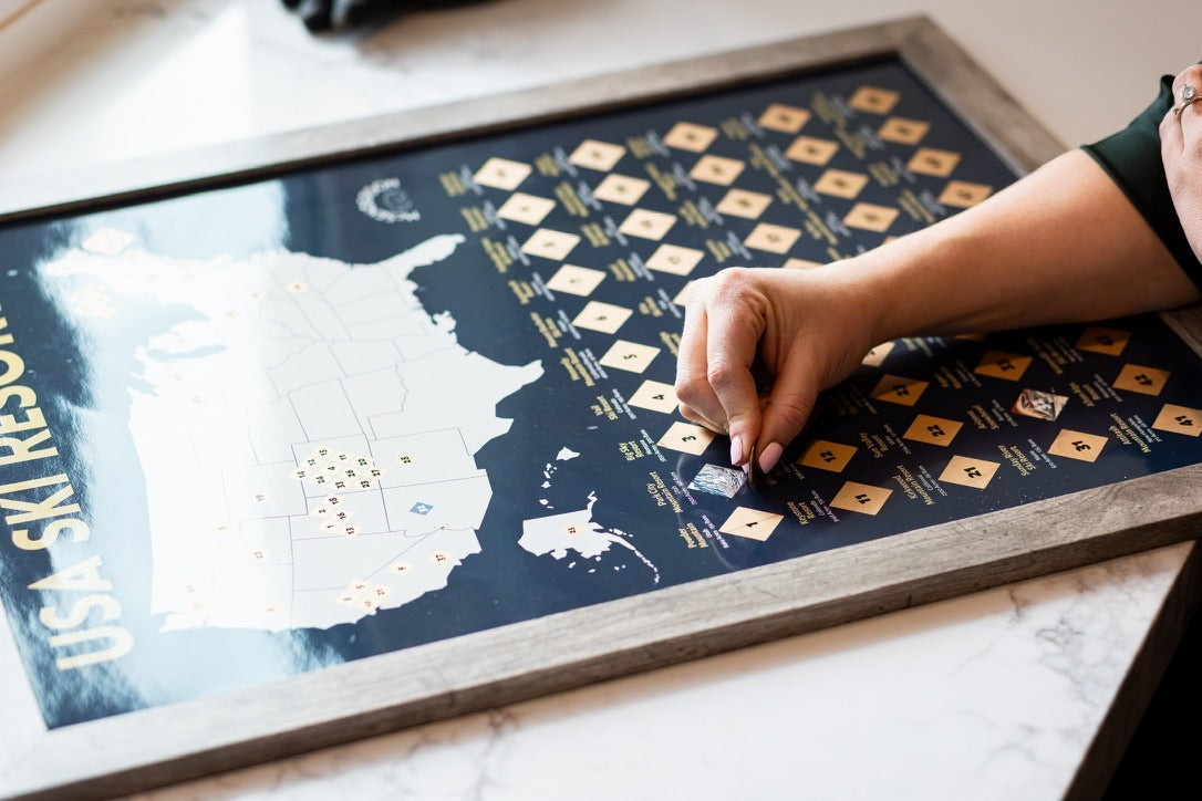 McSenderson Scratch-Off Top 50 USA Ski Resorts Map, vertical orientation, dark blue with gold accents. A hand is scratching off a resort.