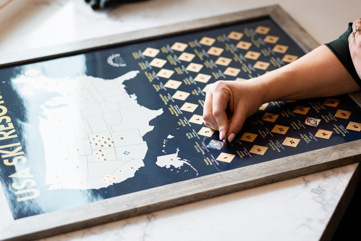 McSenderson Scratch-Off Top 50 USA Ski Resorts Map, vertical orientation, dark blue with gold accents. A hand is scratching off a resort.